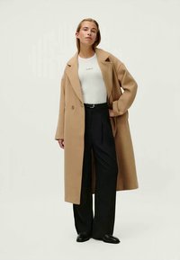 Casaco oversized bege com grandes lapelas, fecho de botão duplo, combinado com uma t-shirt branca ajustada e calças pretas de cintura alta.
