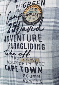 Himmelblaues und weißes kariertes Hemd mit schwarzem Text und einem runden Aufnäher mit Paragliding-Thema, einschließlich der Details "Camp David" und "Kapstadt".