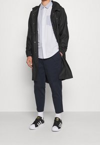 Svart lättviktig jacka över en ljusblå skjorta med knappar och ficka, marinblå capribyxor och svarta sneakers med vita tår.