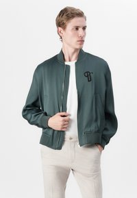 Veste bomber en vert foncé avec une texture douce, fermeture éclair à l'avant, poignets côtelés et un logo noir sur la poitrine. Assortie à un pantalon clair.