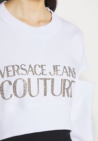 Vit kort sweatshirt med "VERSACE JEANS COUTURE" i metalliska nitar. Rund hals med ribbad struktur och utskärning på axeln.