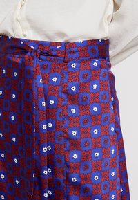 Jupe bleue avec un motif floral rouge et blanc, dotée d'une taille nouée et d'une texture lisse. Le tissu présente un design géométrique avec des carrés.