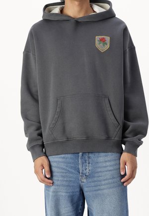 Person trägt einen dunkelgrauen Hoodie mit Fronttasche und kleinem besticktem Schild-Patch mit roter Rose und Text, kombiniert mit blauen Jeans.