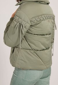Veste verte matelassée avec une capuche structurée, des cordons de serrage sur les manches et un laçage décoratif dans le dos, présentant une texture lisse.
