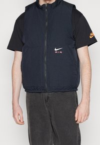 Marinblå nylonväst med dragkedja, ribbad struktur, två framfickor och en vit Nike-logga med "AIR" i rosa. Bärs över en svart t-shirt.