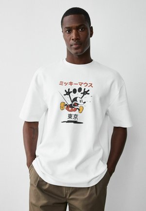 Man wearing white T-shirt with a vintage-style Mickey Mouse graphic and Japanese text, standing against a plain white background.