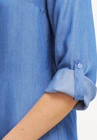 Jasnoniebieska koszula dżinsowa z podwiniętymi rękawami. Posiada detal w postaci guzików oraz subtelną teksturę. Materiał wydaje się gładki, lekki i przewiewny.