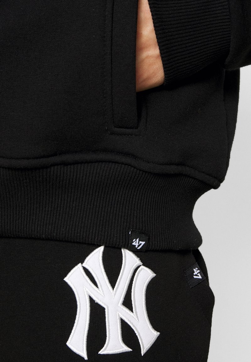 Black sweatshirt featuring a large white embroidered logo, ribbed cuffs, and a visible pocket on the side. Contrasting tags on the hem.