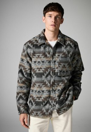 Chaqueta de franela con patrón en gris y marrón, con cuello, cierre de botones, bolsillo frontal y mangas largas, llevada sobre una camisa blanca.
