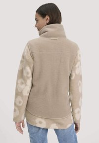 Beige fleece takki, jossa on korkea kaulus; kukkakuosi hihoissa; pehmeä rakenne; kontrastiväriset yksityiskohdat; kaareva helma.