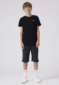 Băiat zâmbitor purtând tricou negru, pantaloni scurți închiși la culoare rulați, șosete albe cu text și adidași albi, stând în picioare pe un fundal simplu, deschis la culoare.