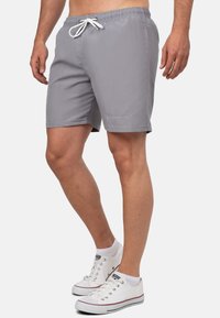 Short de bain gris avec taille élastique à cordon, poches latérales et détail de logo à l'ourlet. Porté avec des baskets blanches et des chaussettes de cheville.