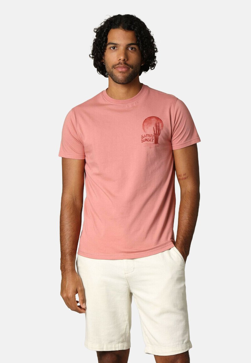 T-shirt en coton rose clair à manches courtes, avec un design graphique rouge représentant un soleil et des cactus étiquetés "SAFARI SUNSET." Un short blanc.