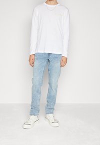 Vit långärmad skjorta med gul "HILFIGER"-logotyp, parat med ljusblå denimjeans och vita sneakers med svarta detaljer.