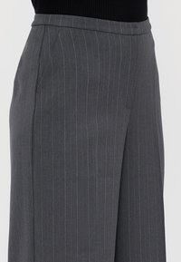 Donkergrijze pinstripe op maat gemaakte broeken met zijzakken, gedragen met een zwarte top, weergegeven van de taille tot halverwege de dijen.