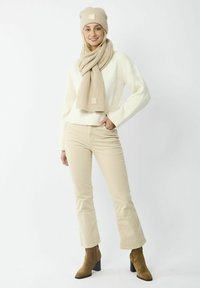 Bonnet en tricot beige et écharpe, pull crème, pantalon évasé beige et bottes à talons marron. Tissu texturé et détail de logo visible sur les accessoires.