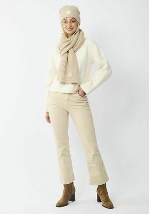 Beige gebreide beanie en sjaal, crèmekleurige trui, beige flared broek en bruine haklaarzen. Gestructureerde stof en zichtbare logodetails op accessoires.