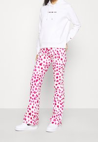 Sudadera blanca con capucha y bolsillo frontal, adornada con un logo negro, combinada con pantalones rosas y blancos que presentan un patrón abstracto de manchas y un corte acampanado.