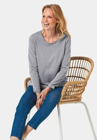 Graues Langarmshirt mit rundem Halsausschnitt, kombiniert mit dunkelblauen Jeans, sitzend auf einem geflochtenen Rattanstuhl mit weißen Metallbeinen.