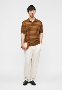 Άνδρας που φοράει καφέ ριγωτό κοντομάνικο πόλο, λευκό παντελόνι με φαρδιά μπατζάκια, μαύρα παπούτσια, κρατώντας μικρή καφέ τσάντα, στέκεται μπροστά σε λευκό φόντο.