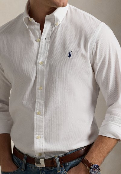 Κοντομάνικο άσπρο πουκάμισο με υφή ύφανσης, που διαθέτει μπλε λογότυπο στο στήθος, σε συνδυασμό με καφέ δερμάτινη ζώνη και ρολόι.