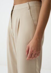 Pantaloni beige a pieghe con chiusura a bottone e zip, dotati di tasche laterali e un tessuto liscio e testurizzato. Inquadratura ravvicinata sulla vita e sulla mano.