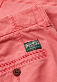 Tela de mezclilla rosa con una superficie texturada, que presenta una etiqueta verde de "Superdry" y un detalle de botón marrón. Costura visible.
