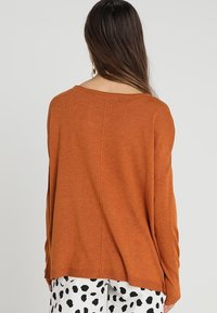 Lös, långärmad tröja i rostorange, stickad struktur, vid halsringning och rak nederkant, bärs över vita byxor med svart prickmönster.