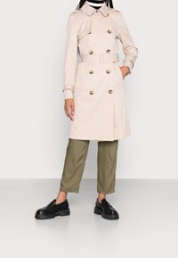 Ljusbeige trenchcoat med dubbelradiga guldfärgade knappar, bälte i midjan och två framfickor, kombinerad med olivgröna byxor och svarta skor.
