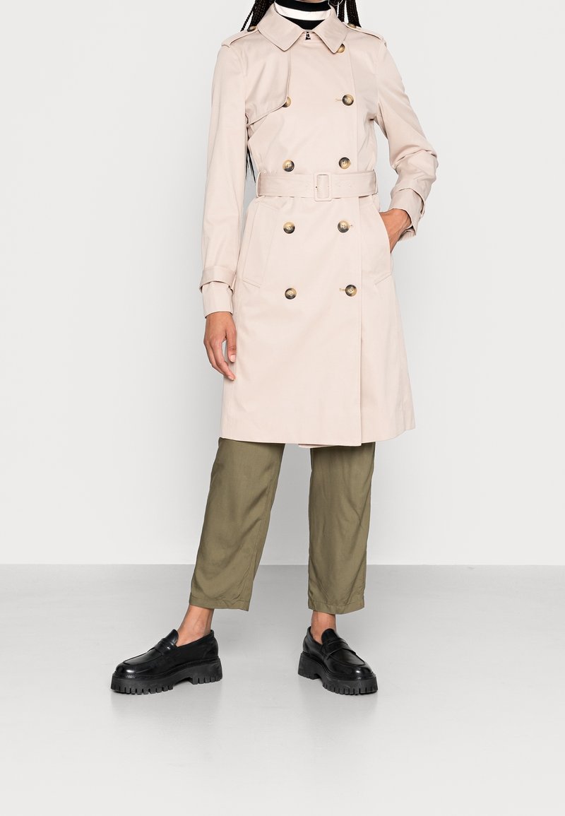 Ljusbeige trenchcoat med dubbelradiga guldfärgade knappar, bälte i midjan och två framfickor, kombinerad med olivgröna byxor och svarta skor.