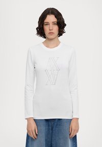 T-shirt bianco a maniche lunghe in cotone con scollatura rotonda. Presenta un design di logo geometrico in borchie argentate sulla parte frontale. Gonna in denim mostrata.