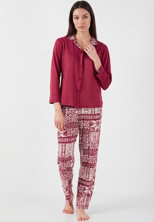 Naine kannab burgundia nuppu nööpidega pika varrukaga ülaosa, millel on mustriline krae ning ühesuguste mustriliste trennipükstega, seisab paljajalu valgel põrandal.