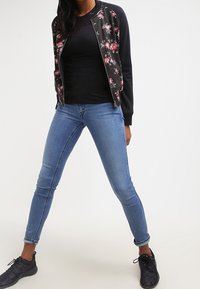 Giacca bomber nera con stampa floreale, top fitted nero, jeans skinny blu e scarpe da ginnastica nere. Look casual a strati con colori in contrasto.