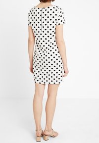 Dlouhý rukáv, přiléhavé šaty v bílé barvě s velkými černými puntíky, s obousměrným zipem na zádech a lemem nad koleny, spárované s nahými sandály.