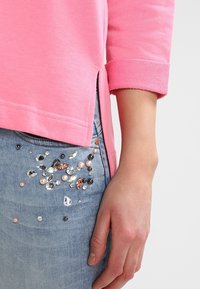 Haut en coton rose avec manches retroussées et fentes latérales. Jean en denim bleu clair orné de diverses perles colorées et de sequins.