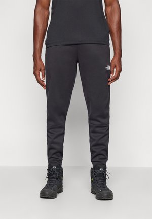 Mężczyzna noszący czarne, dopasowane spodnie dresowe z logo The North Face na lewym udzie oraz czarne buty trekkingowe.