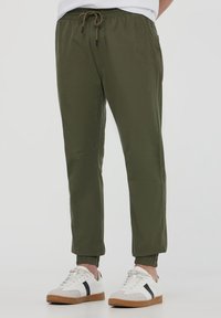 Pantaloni jogger elasticizzati verde oliva con vita elasticizzata e coulisse, caratterizzati da gambe affusolate e polsini a costine. Abbinati a sneakers bianche.