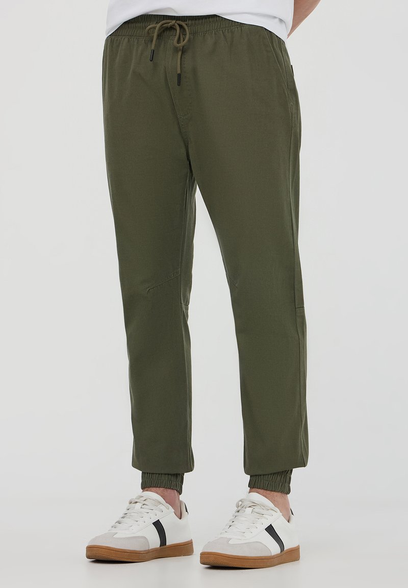 Pantaloni jogger elasticizzati verde oliva con vita elasticizzata e coulisse, caratterizzati da gambe affusolate e polsini a costine. Abbinati a sneakers bianche.