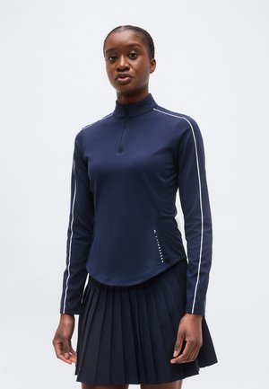 Γυναίκα που φορά μπλε σκούρο τοπ με φερμουάρ μακριάς μανσέτας και λευκό περίγραμμα, μαζί με πλισέ μπλε σκούρο φούστα, στέκεται μπροστά σε μονόχρωμο φόντο.