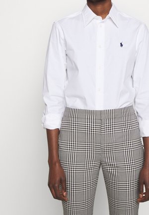 Personne portant une chemise blanche à manches longues avec un petit logo brodé et un pantalon à carreaux noir et blanc.