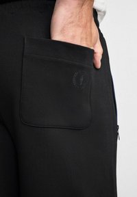 Czarne dresy z tylną kieszonką z teksturowanym logo, suwakiem po bokach oraz gładkim, lekkim materiałem.