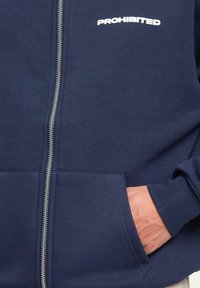 Navy blue zip-up hoodie with white "PROHIBITED" text on chest, featuring front pockets and ribbed cuffs, hand inside right pocket.