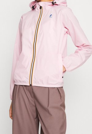 Veste à capuche rose clair avec fermeture éclair rayée et logo sur la poitrine, portée avec un pantalon marron ample.