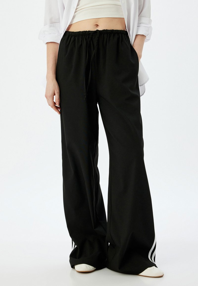 Pantalones anchos negros con cintura elástica, cordón y rayas laterales blancas. Hechos de una tela suave con una textura lisa.