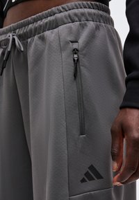 Κοντινό πλάνο γκρι φόρμας Adidas με ελαστική μέση, τσέπη με φερμουάρ και μαύρο λογότυπο κοντά στην τσέπη, φορεμένη από ένα άτομο με σκουρόχρωμο δέρμα.