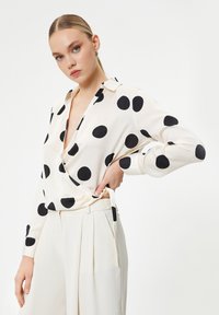 Blusa bianca a maniche lunghe con grandi pois neri, caratterizzata da un design avvolgente e un colletto, abbinata a pantaloni plissettati color crema chiaro.