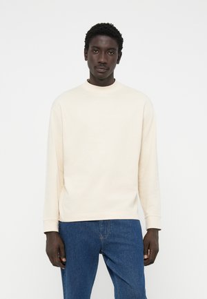 Suéter largo de manga larga en beige claro con un cuello alto, hecho de un material suave, combinado con jeans azules. Diseño simple, con detalles mínimos.