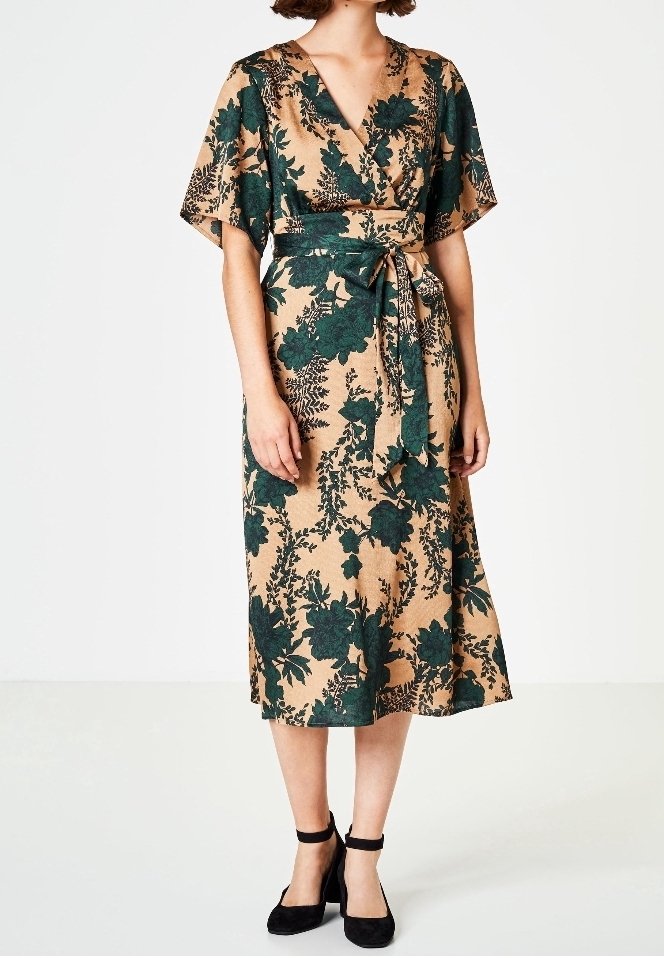 Robe portefeuille beige avec imprimé floral vert foncé, manches courtes volantées, ceinture à la taille, portée avec des talons noirs à bride cheville sur un mannequin.