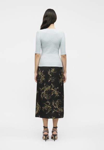 Γυναίκα με μακριά σκούρα μαλλιά που φοράει ανοιχτόχρωμο εφαρμοστό τοπ και μαύρη midi φούστα με πράσινα και ροζ λουλουδάτα σχέδια, όρθια με την πλάτη προς τα εμπρός.