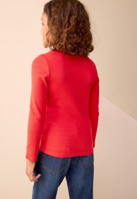 Red ribbed long-sleeve shirt with a round neckline, paired with blue denim jeans. Texture is prominent, showing a casual fit.